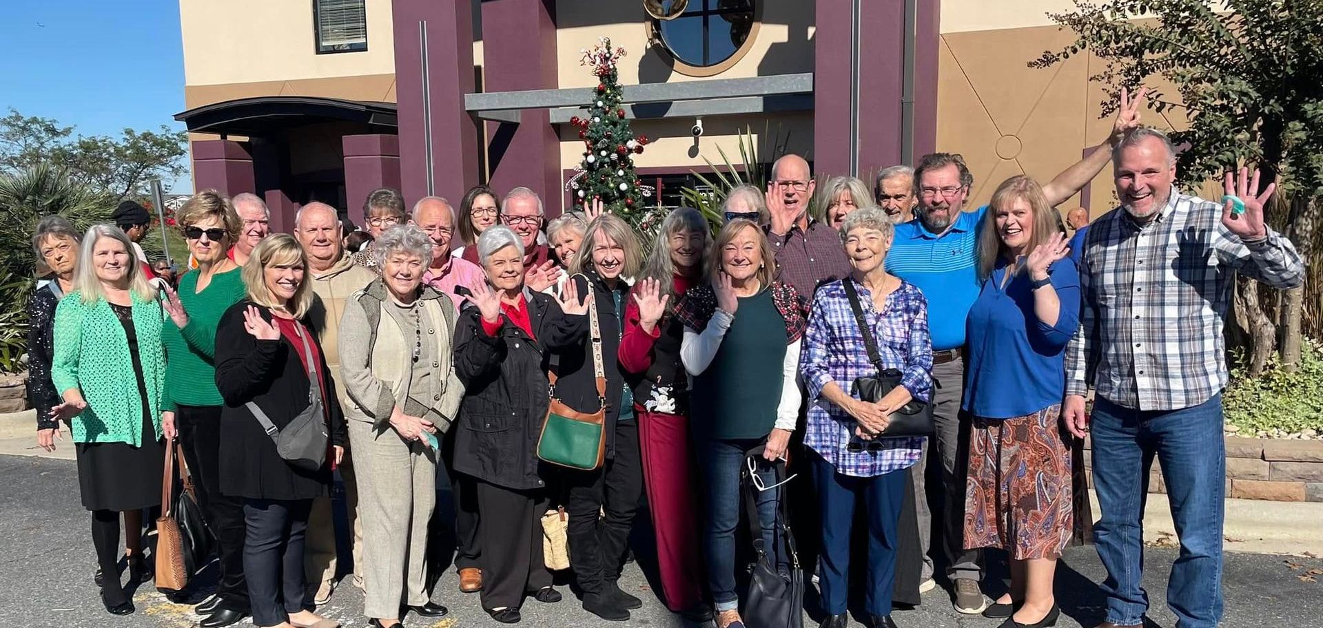 A group of mostly older adults outside a building, waving and smiling. Building has purple accents.
