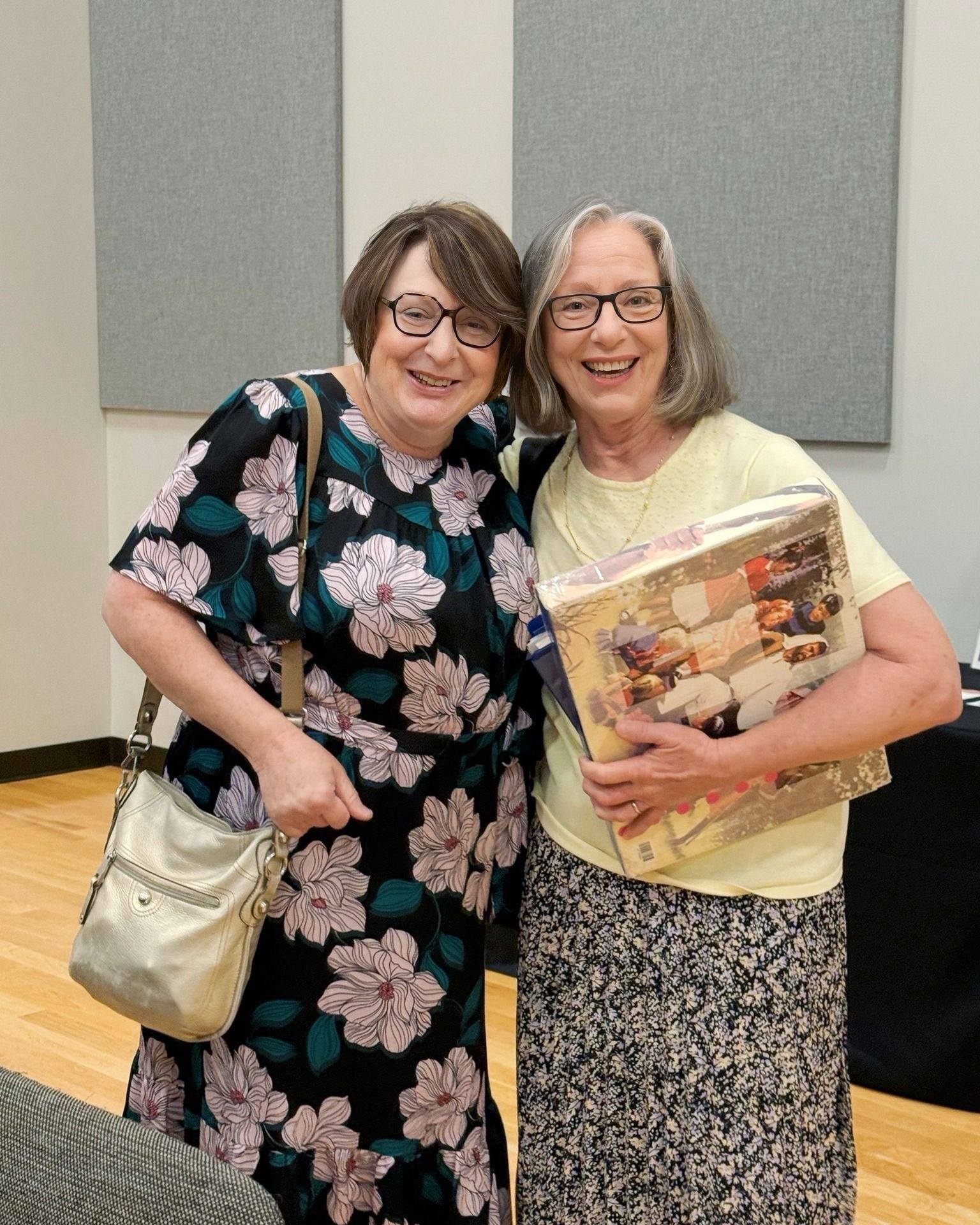 Two women smiling and posing indoors, one in a floral dress, the other with a book in hand.