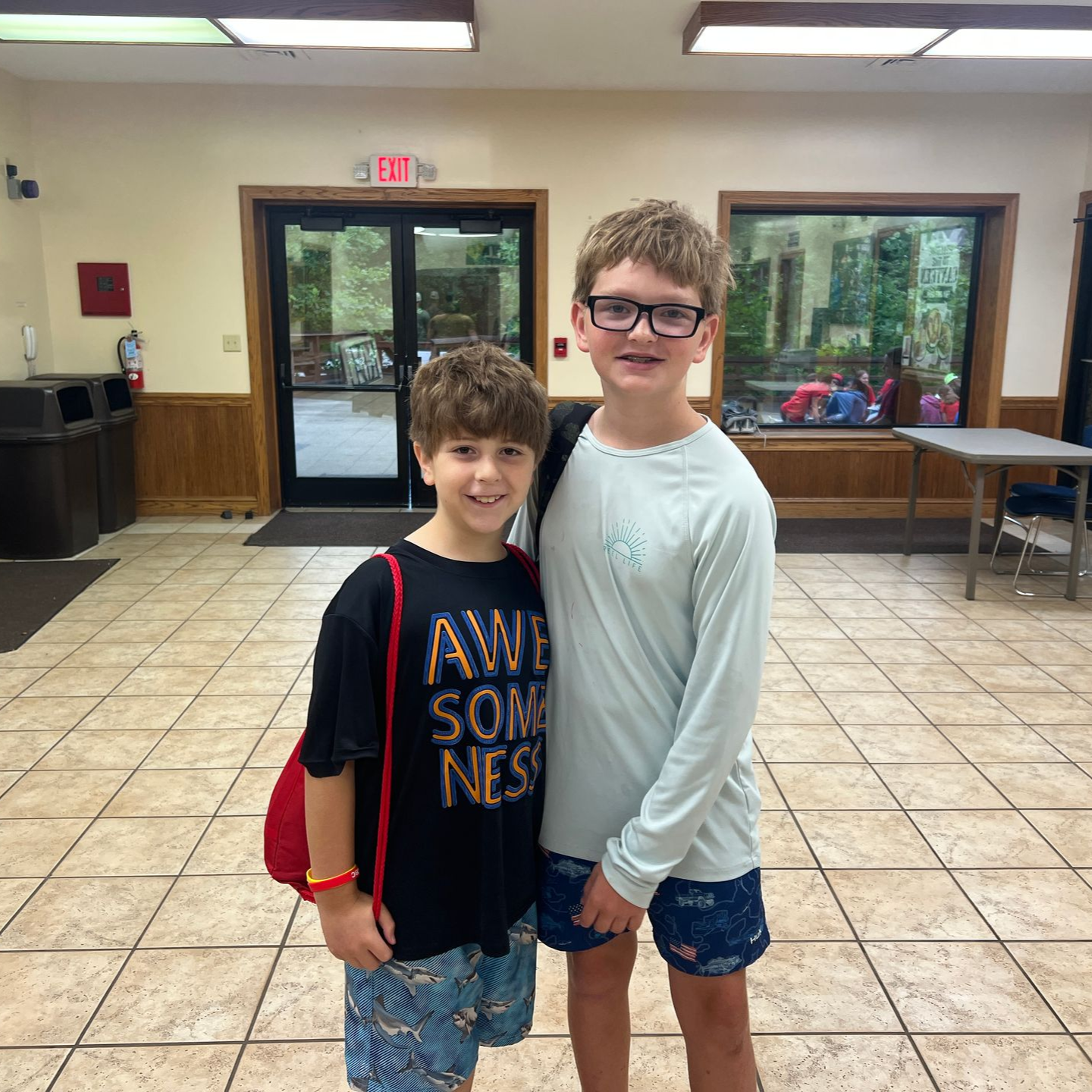 Two smiling boys looking at the camera at a table. One wears an orange plaid shirt. Other, blue plaid.