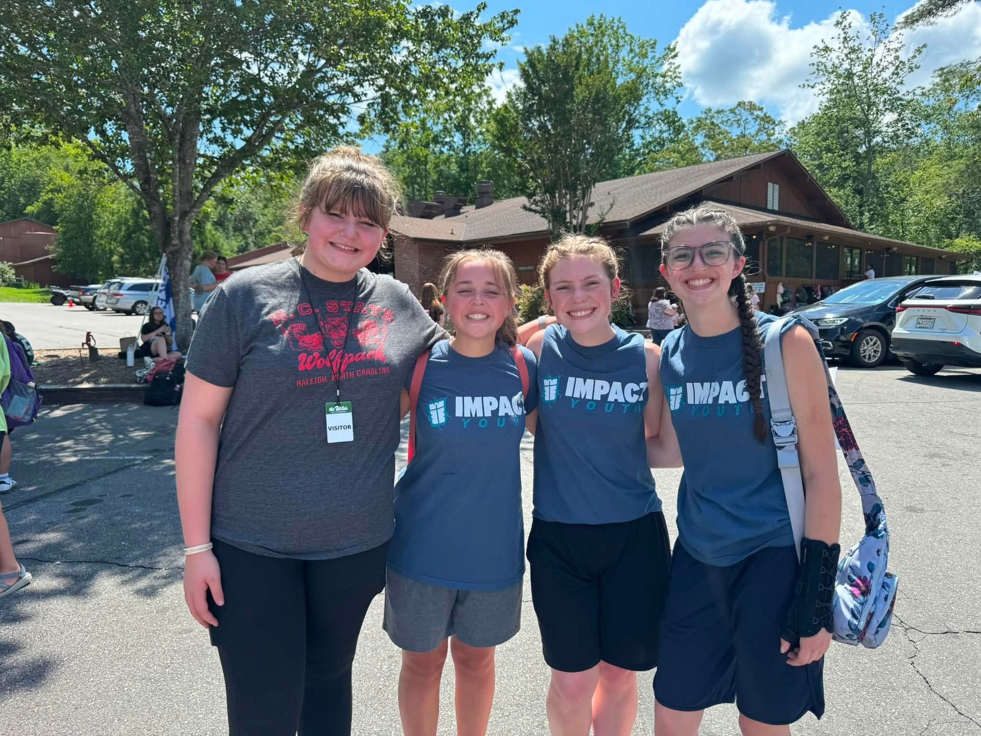 Five young people smile at a table outdoors. They wear lanyards, near trees and a building.