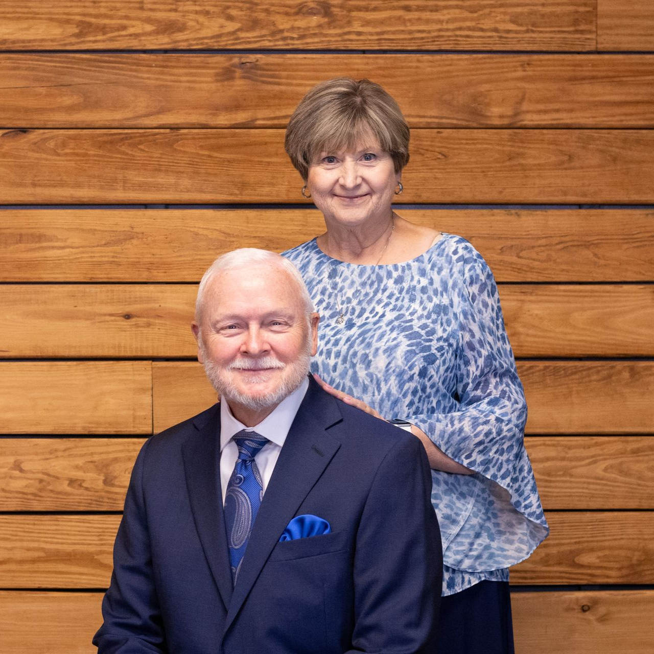 An older couple stands in front of a building. The woman wears blue and patterned skirt, man blue shirt and pants.