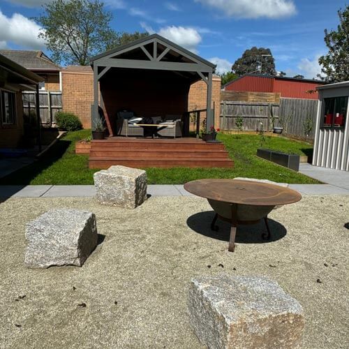 Backyard patio with gravel seating area, stone blocks, round table, and a covered deck with stairs and lawn.