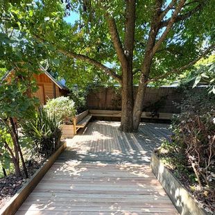 Wooden garden walkway leading to a shaded patio beneath a large tree, bordered by raised planters and a fence