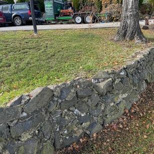 Stone retaining wall beside a grassy yard with parked vehicles and a large tree in the background
