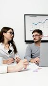 Three coworkers discussing charts in a meeting room with a wall-mounted graph display and laptop.