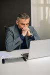 Man in a gray suit thinking at a desk with a laptop and notebook in a bright office