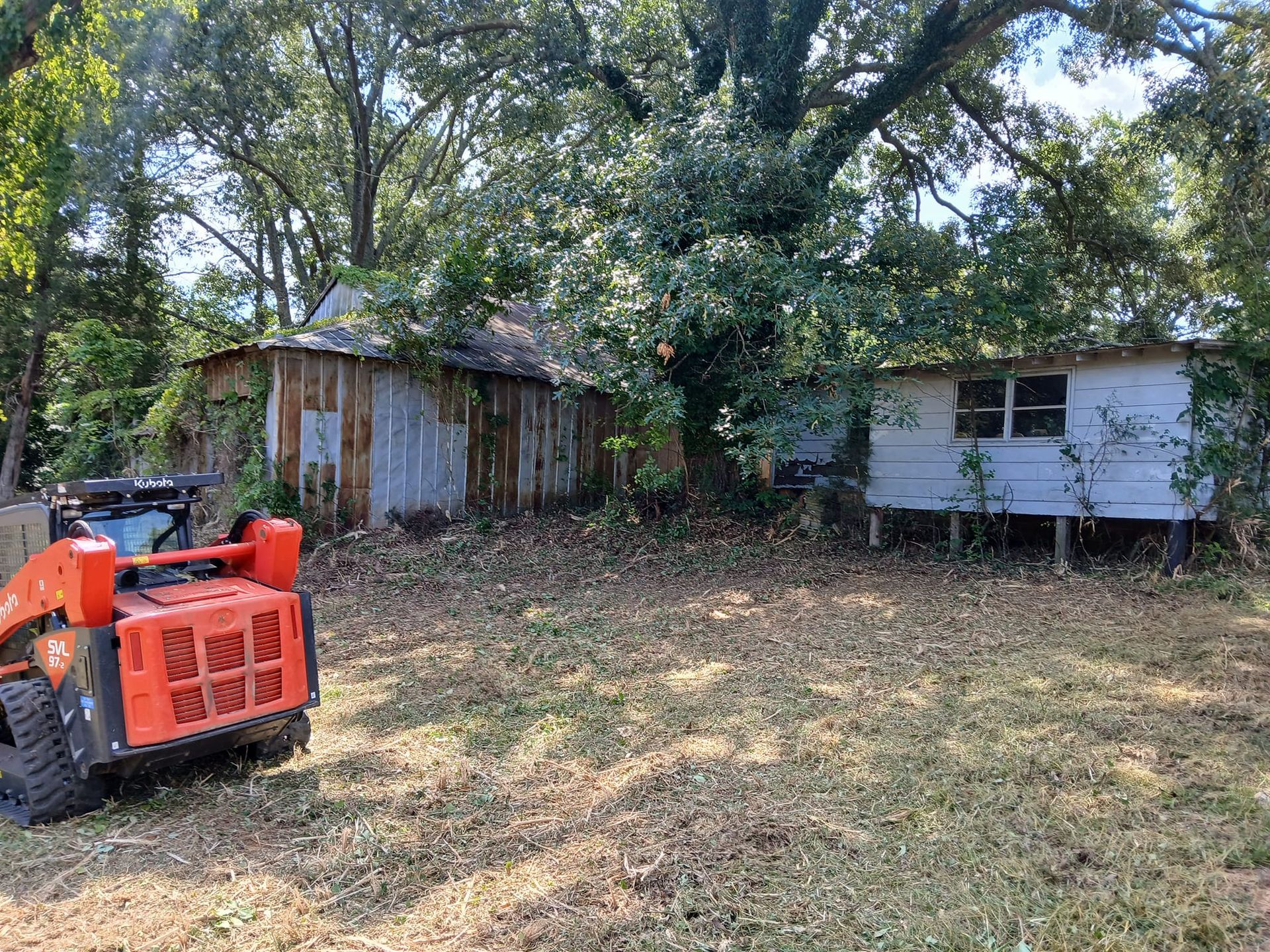 Orange tractor in a wooded yard beside two small weathered buildings.