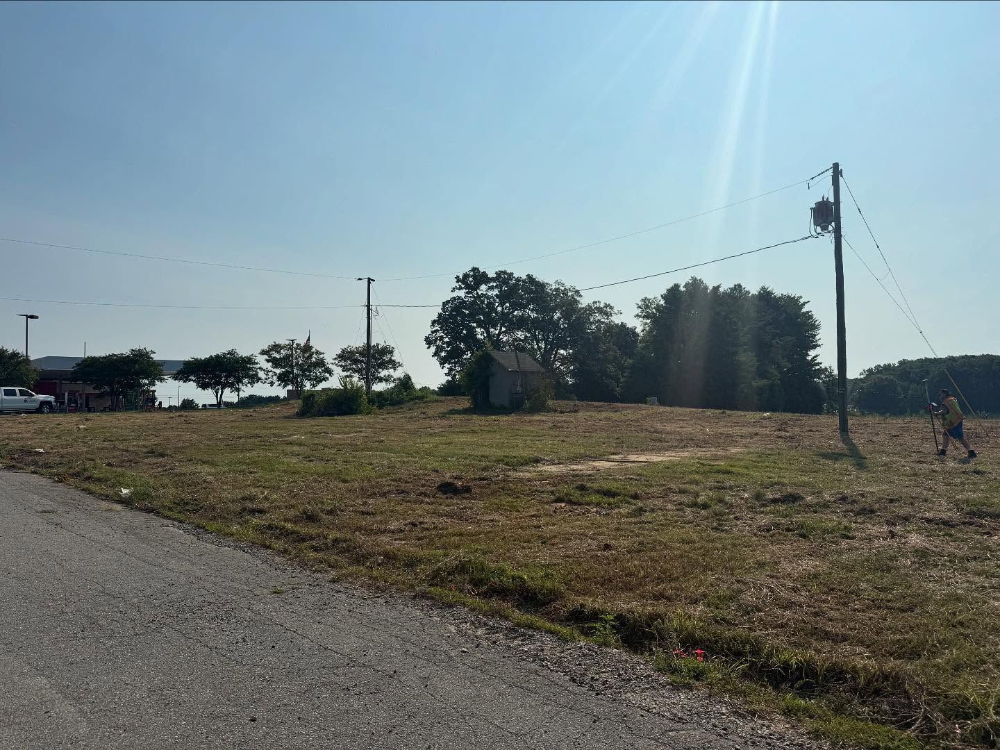 Sunny rural roadside with dry grass, scattered trees, utility poles, and a paved road in the foreground