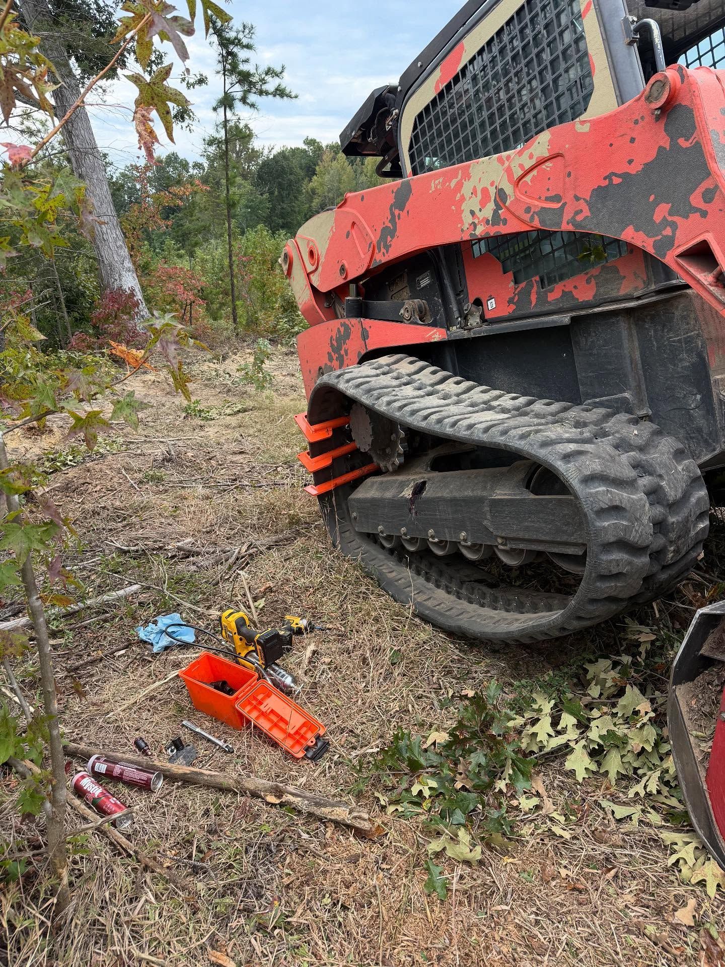 Red skid-steer track beside a wooded trail, with an orange chainsaw on the ground.