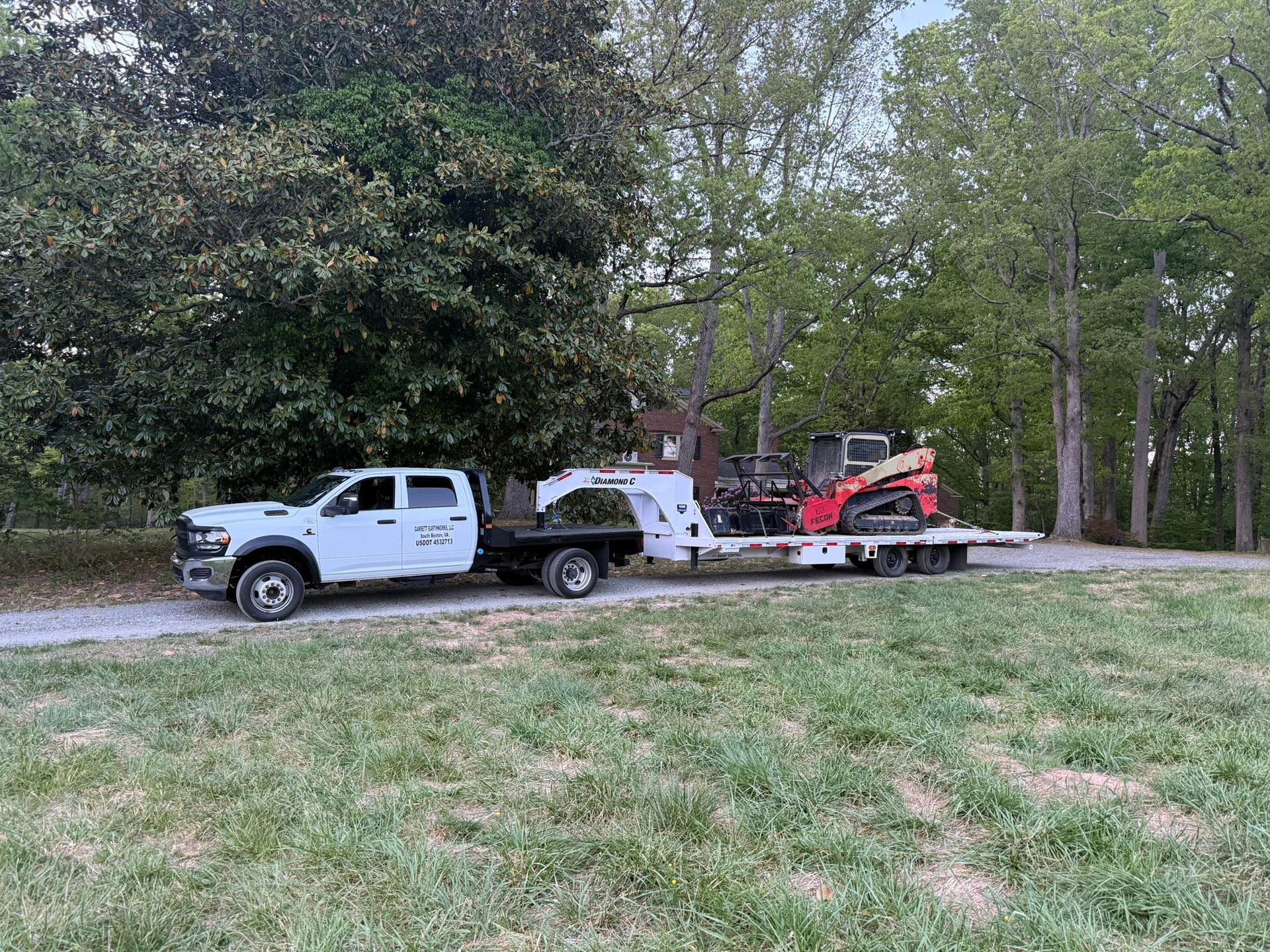 White tow truck hauling a white truck and red skid steer on a flatbed beside a wooded roadside