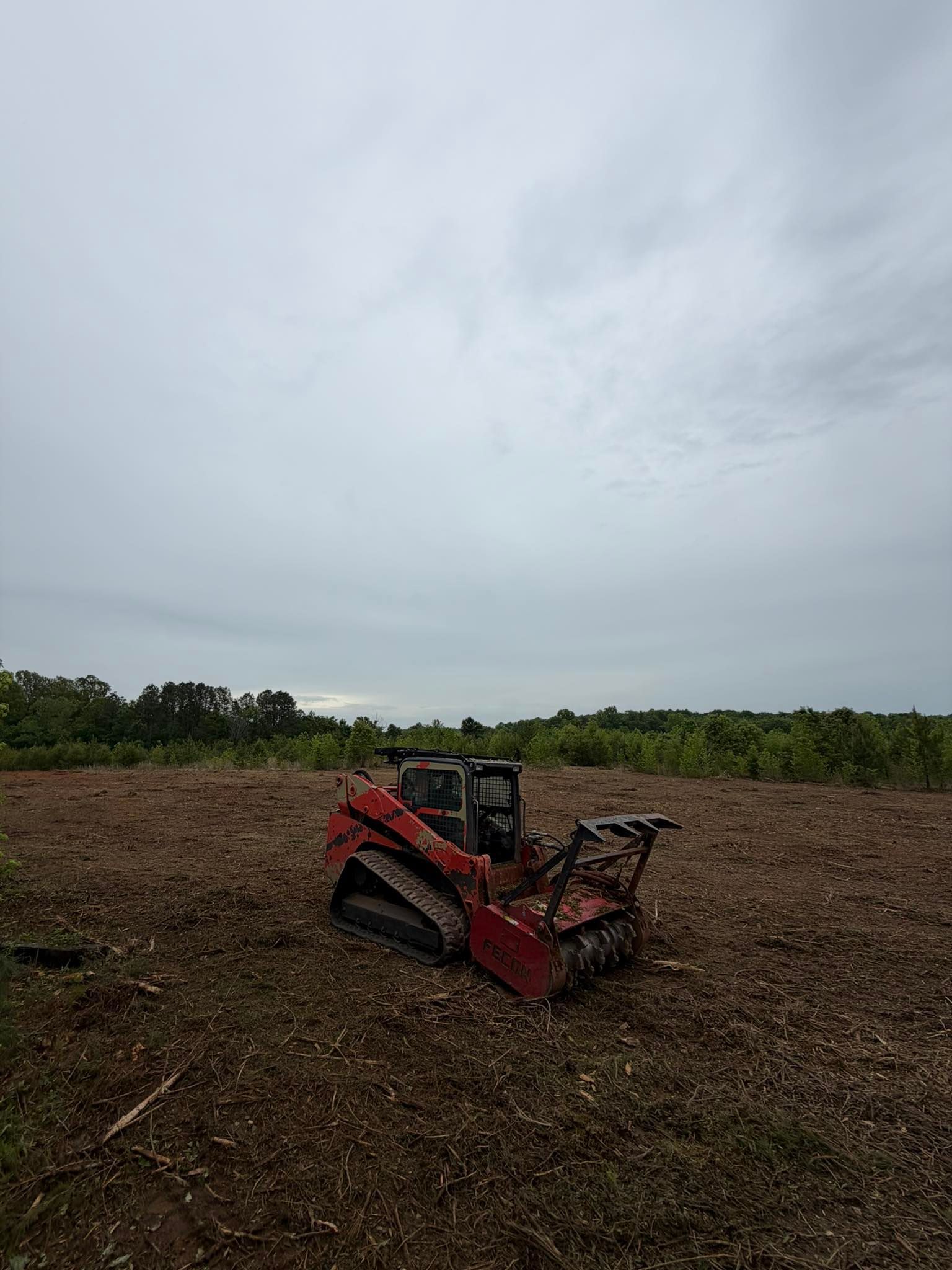 Red farm machine in a muddy field under a cloudy sky, with trees along the horizon.