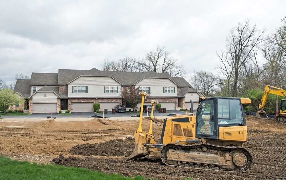 Yellow excavators working on a dirt lot in front of suburban houses under a cloudy sky