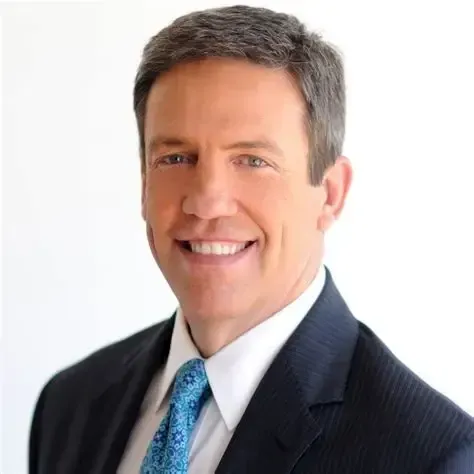 Man in suit smiling, with blue patterned tie, against a white background.