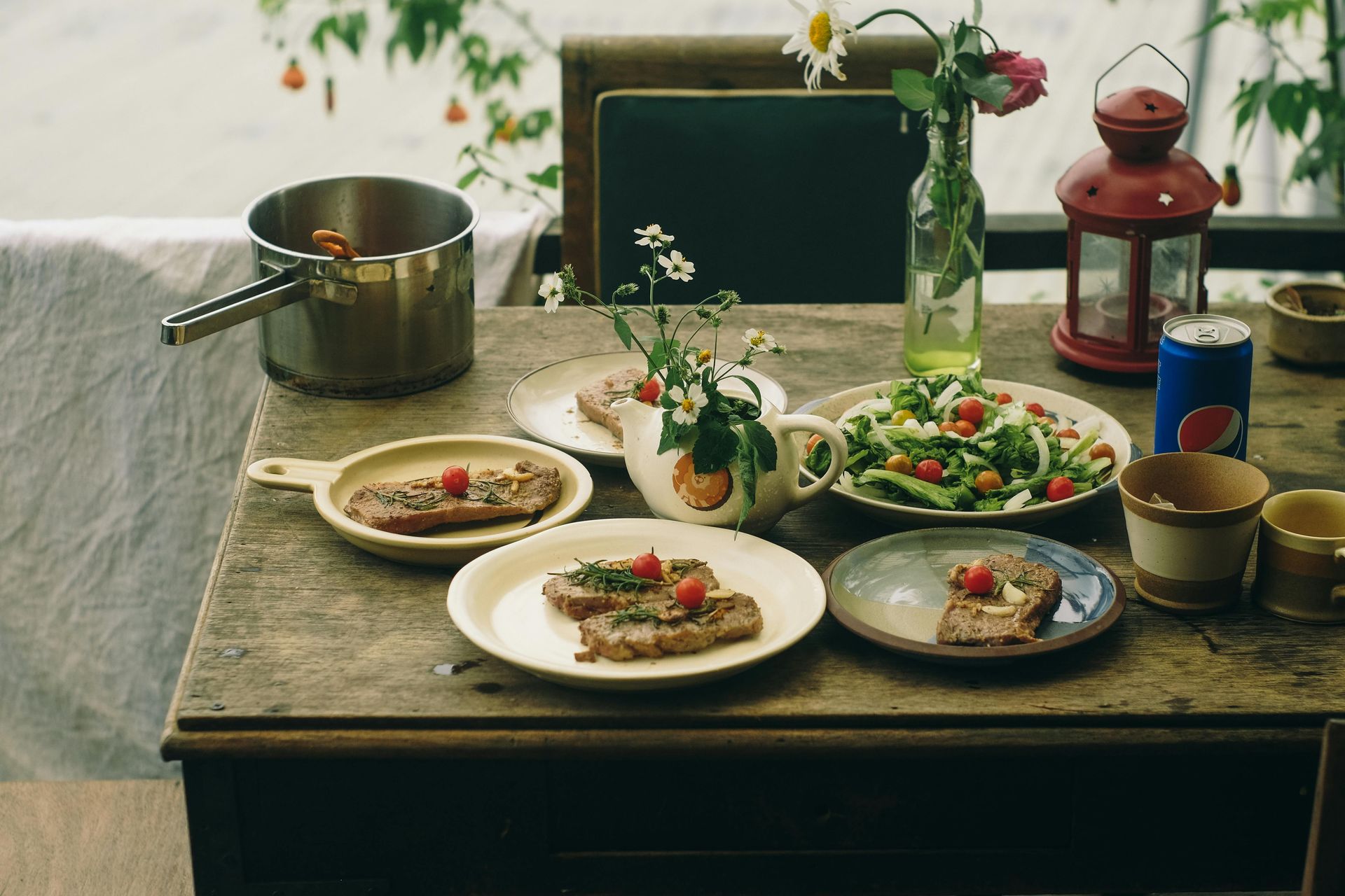 Table set with a meal including plates of meat, salad, and a Pepsi can, flowers, and a red lantern.