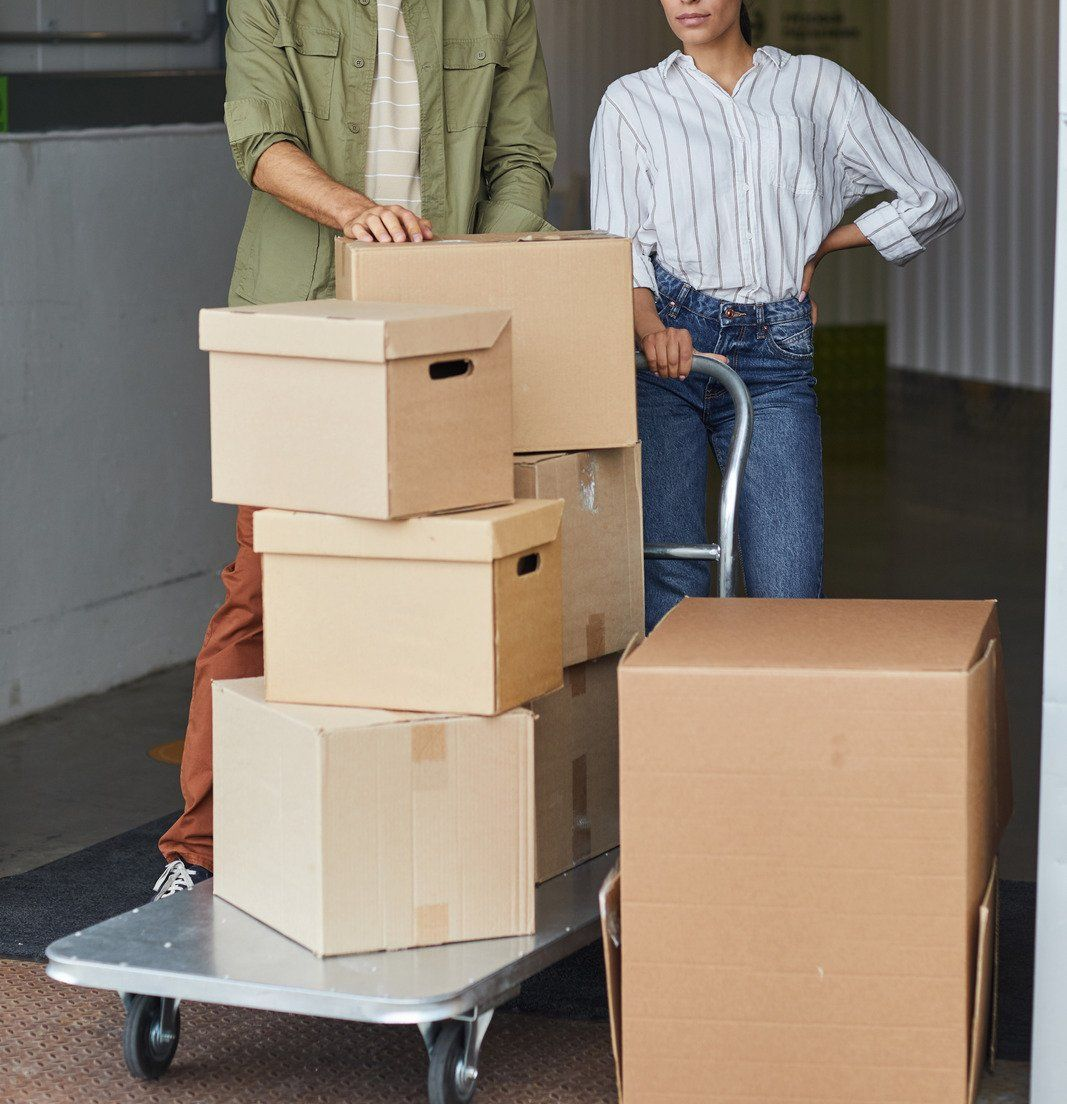 Two people moving stacked cardboard boxes on a hand truck at a loading area
