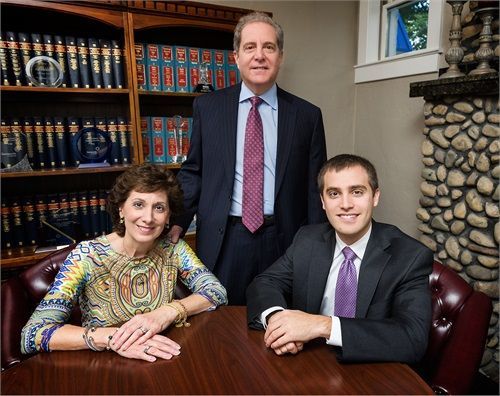 Three people, two seated and one standing, in an office setting. They are smiling, presumably lawyers. Bookshelves and a stone fireplace are visible.