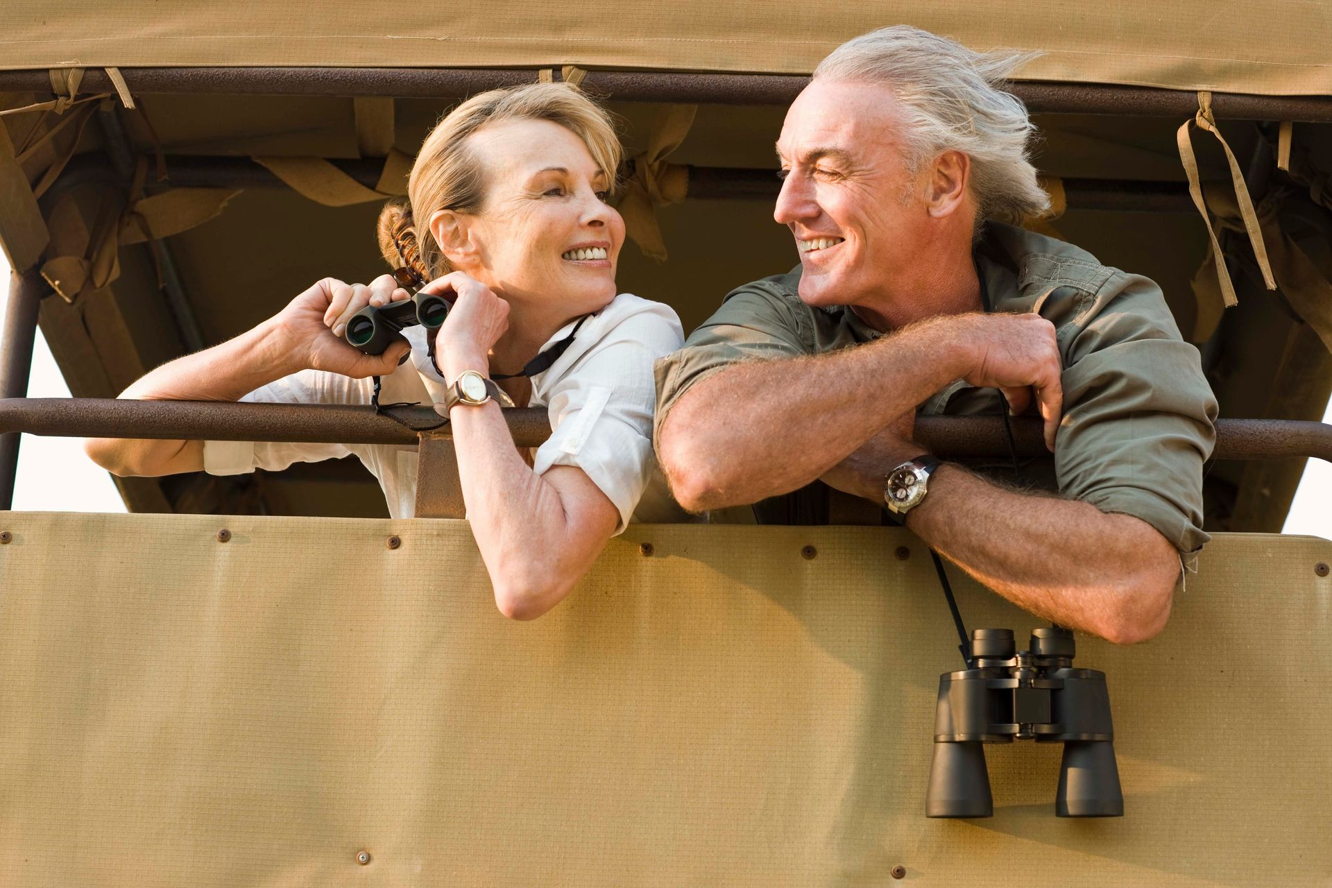 A smiling older couple in a safari vehicle, looking at each other. Woman holds binoculars; man has his arms crossed.
