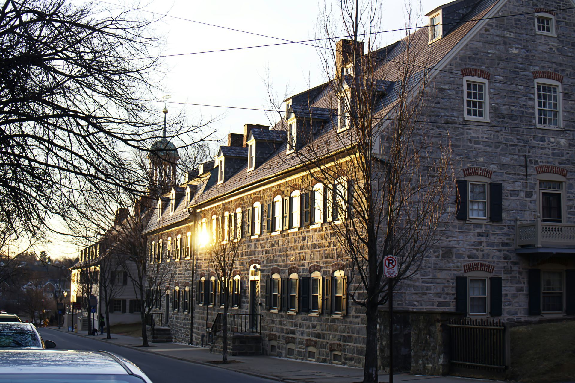 Stone building with multiple windows and dormers, lit by the sun. A street with parked cars and bare trees is in the foreground.