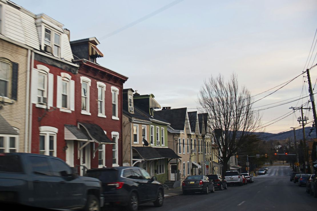 Row houses with various colors and dormer windows line a street with parked cars on both sides.