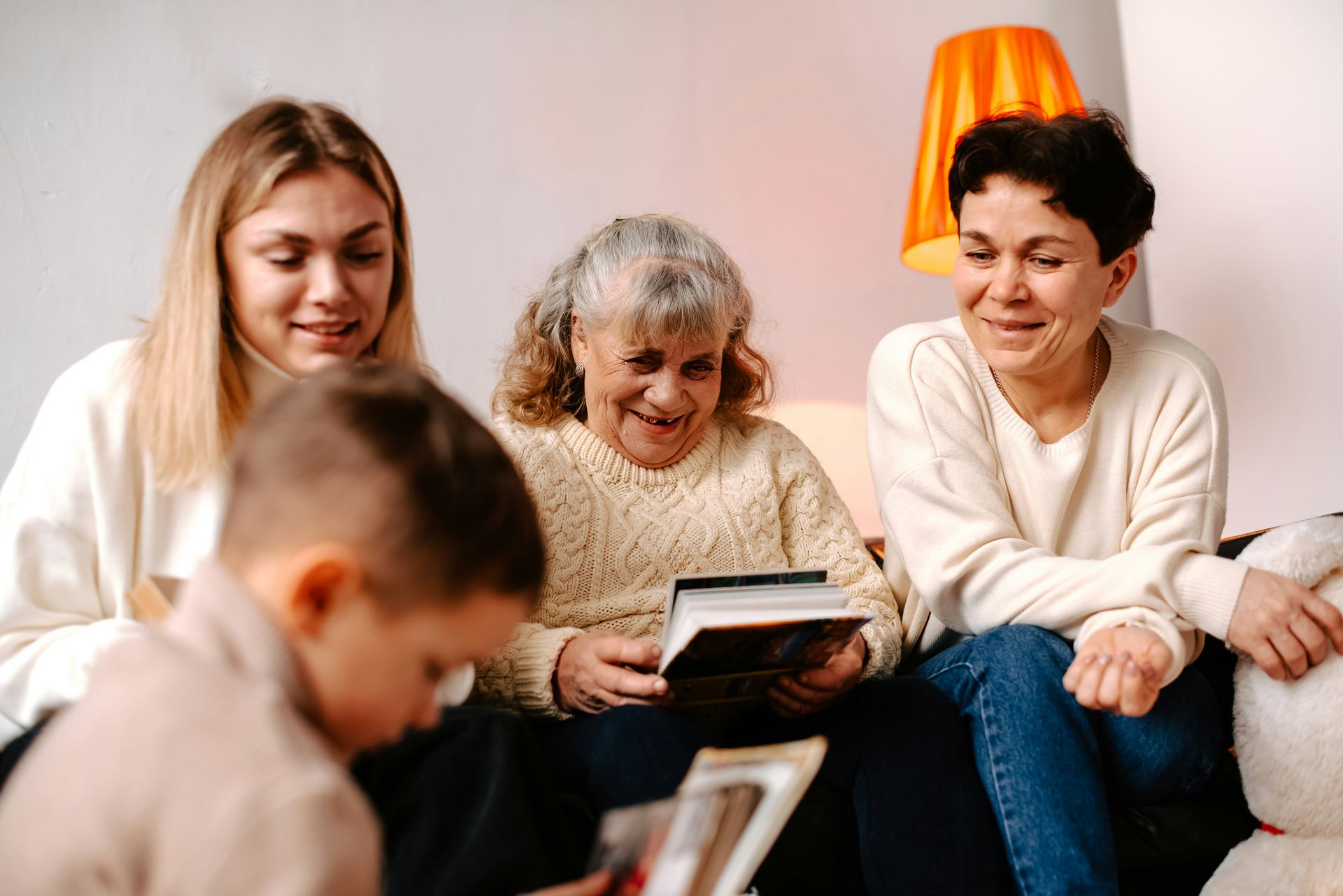 Four generations looking at photo albums: a child, young woman, older woman, and woman smiling on a couch indoors.