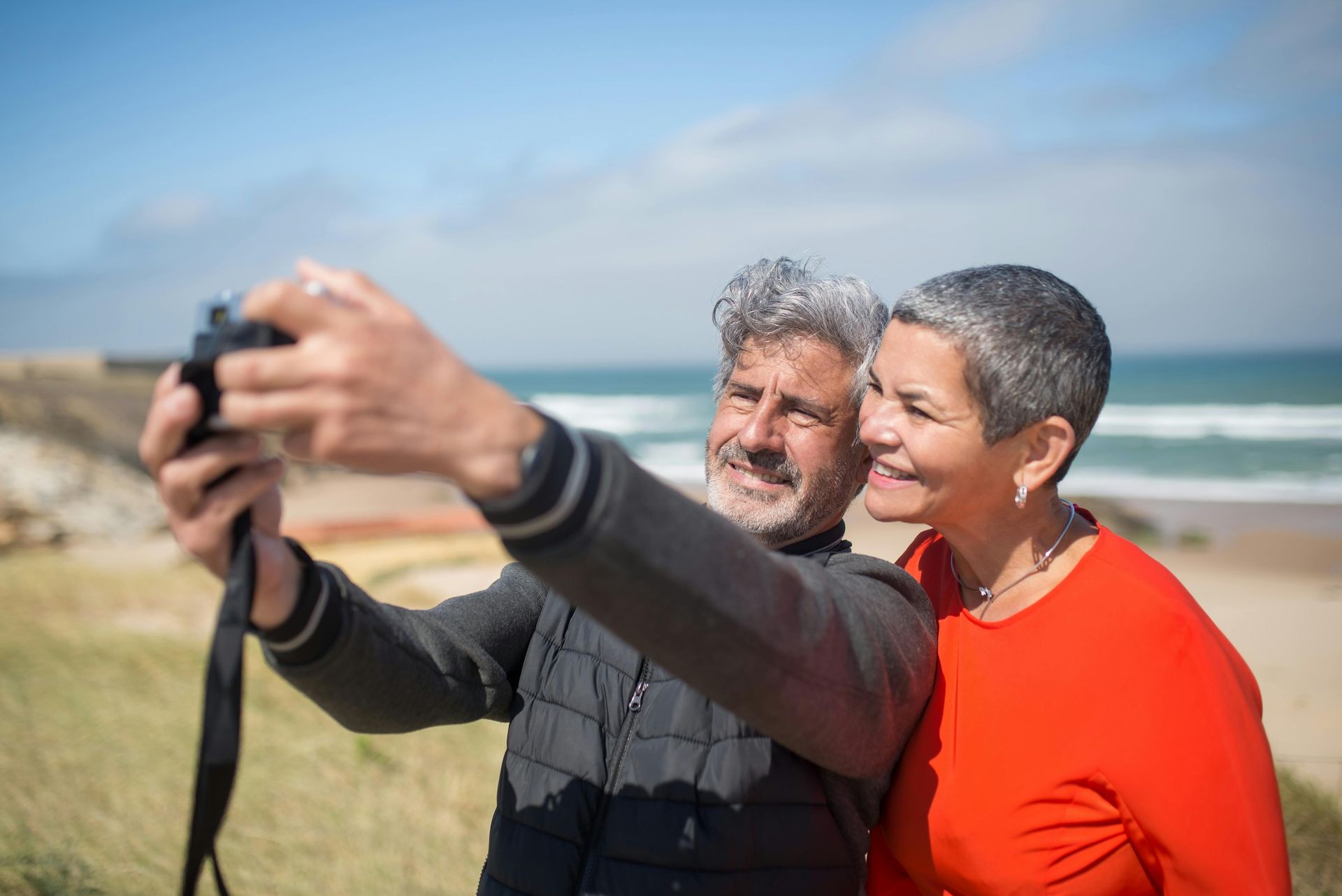 Older couple smiling, taking a selfie with a camera on a beach. The woman wears orange, the man has gray hair.