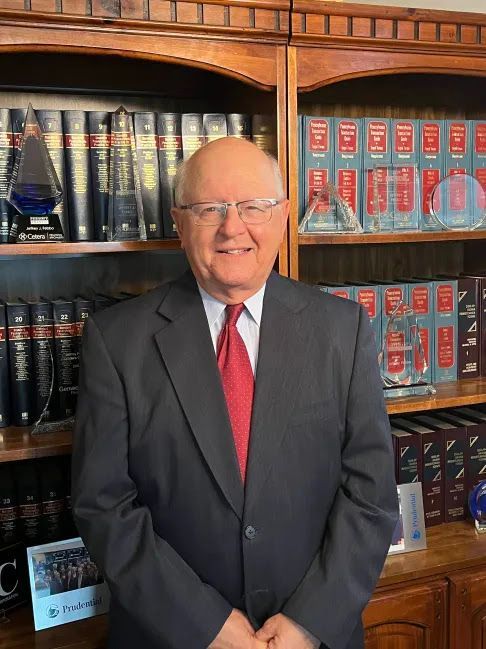 Bald man in a dark suit and red tie smiles in front of a wooden bookshelf filled with law books and awards.