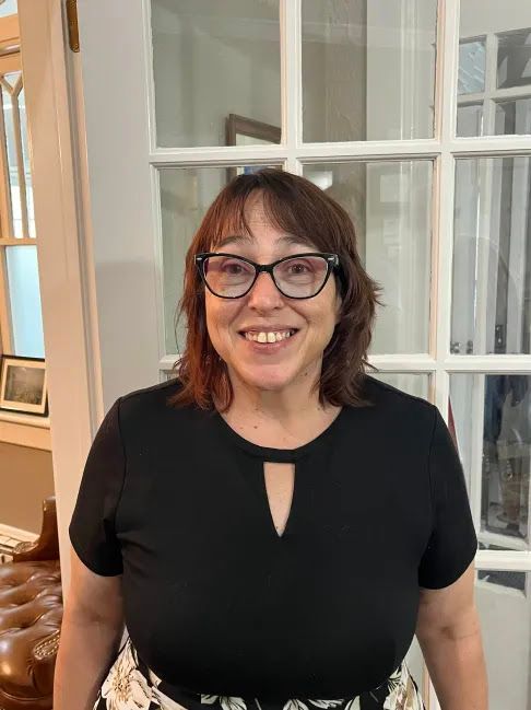 Woman with glasses smiles, wearing a black top and patterned skirt, standing in front of a glass-paneled door.