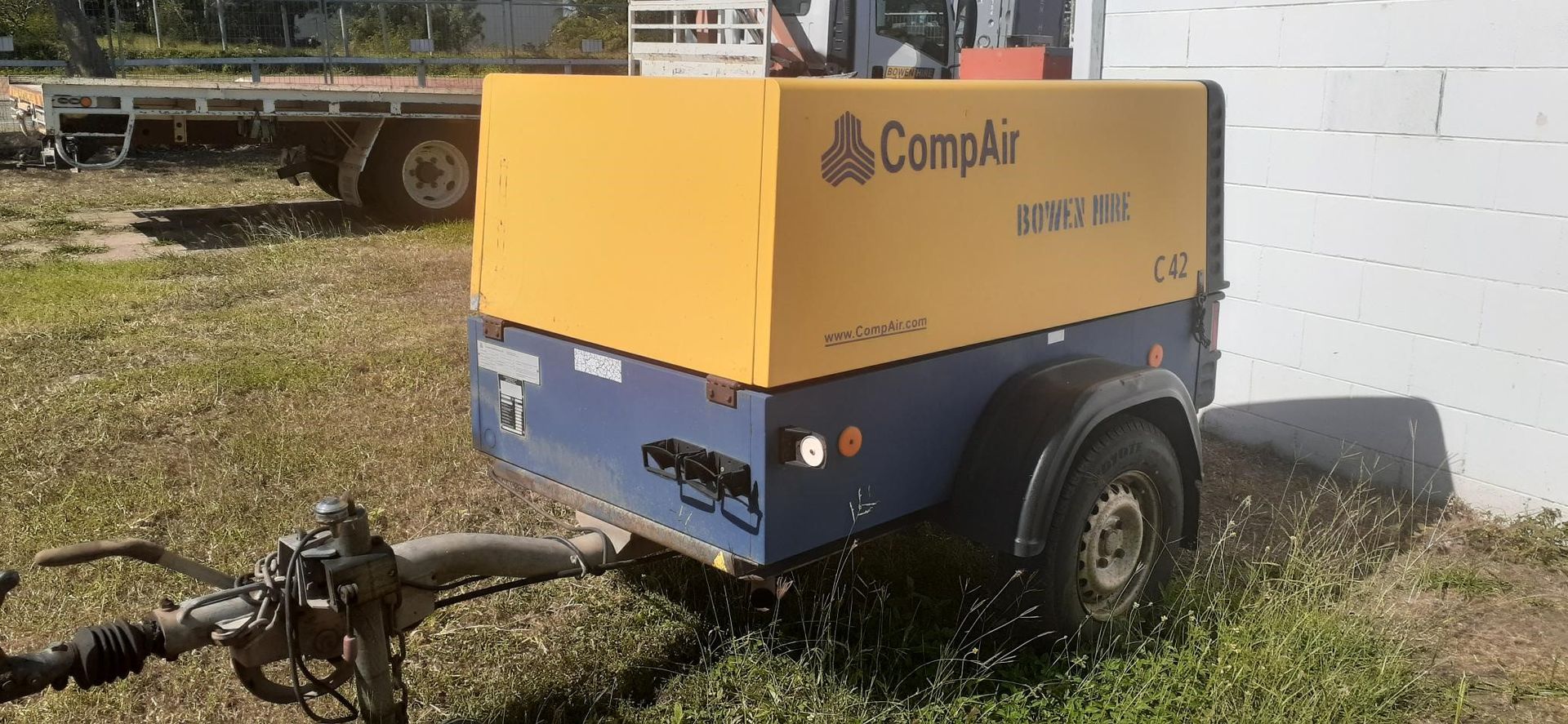Yellow and blue CompAir portable air compressor trailer parked on gravel by a white wall — Bowen Hire in Bowen, QLD
