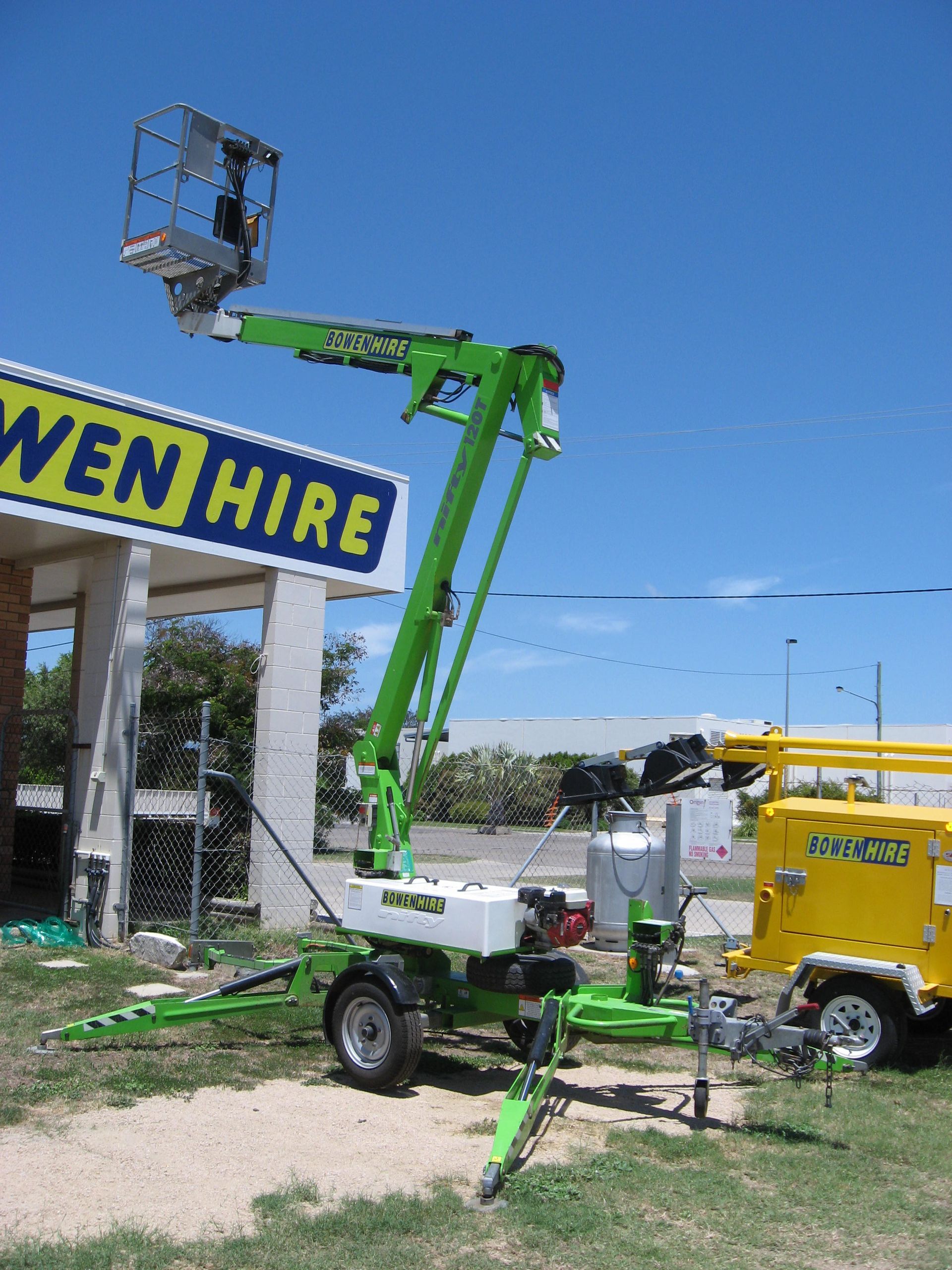 Green boom lift with worker basket beside a yellow WEM Hire truck under a blue sky — Bowen Hire in Bowen, QLD 
