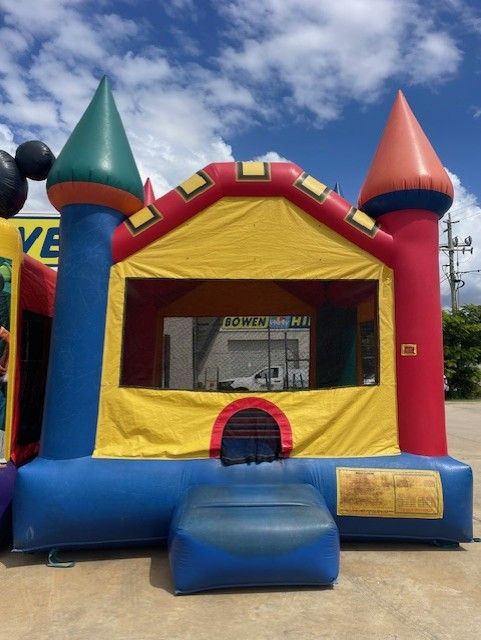 Colorful inflatable bounce house with yellow walls, red roof, blue base, and cone towers outdoors under a cloudy sky - Bowen Hire in Bowen, QLD