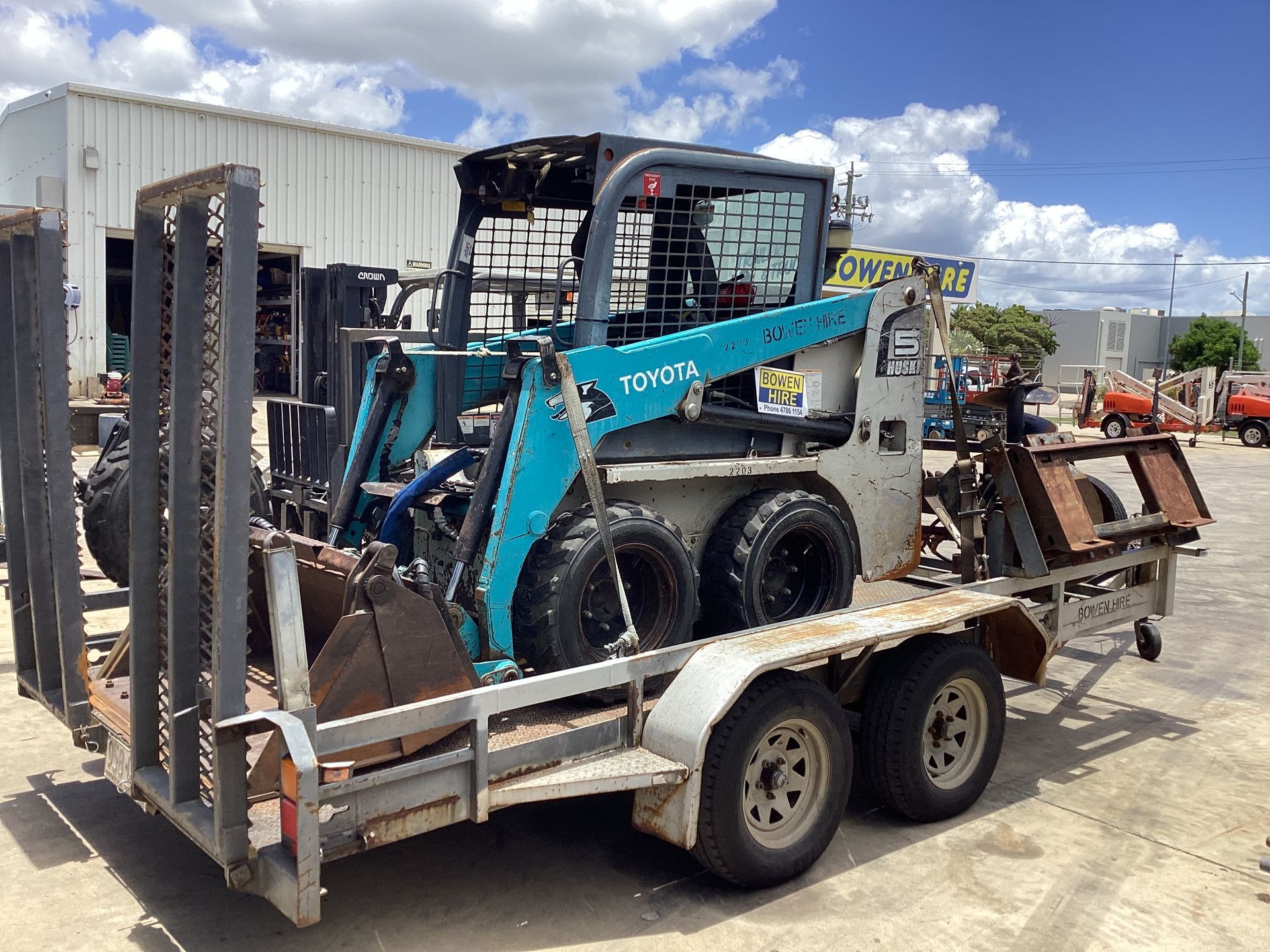 Blue Skid-steer Loader on a Trailer in an Industrial Yard, With Other Equipment Nearby — Bowen Hire in Bowen, QLD