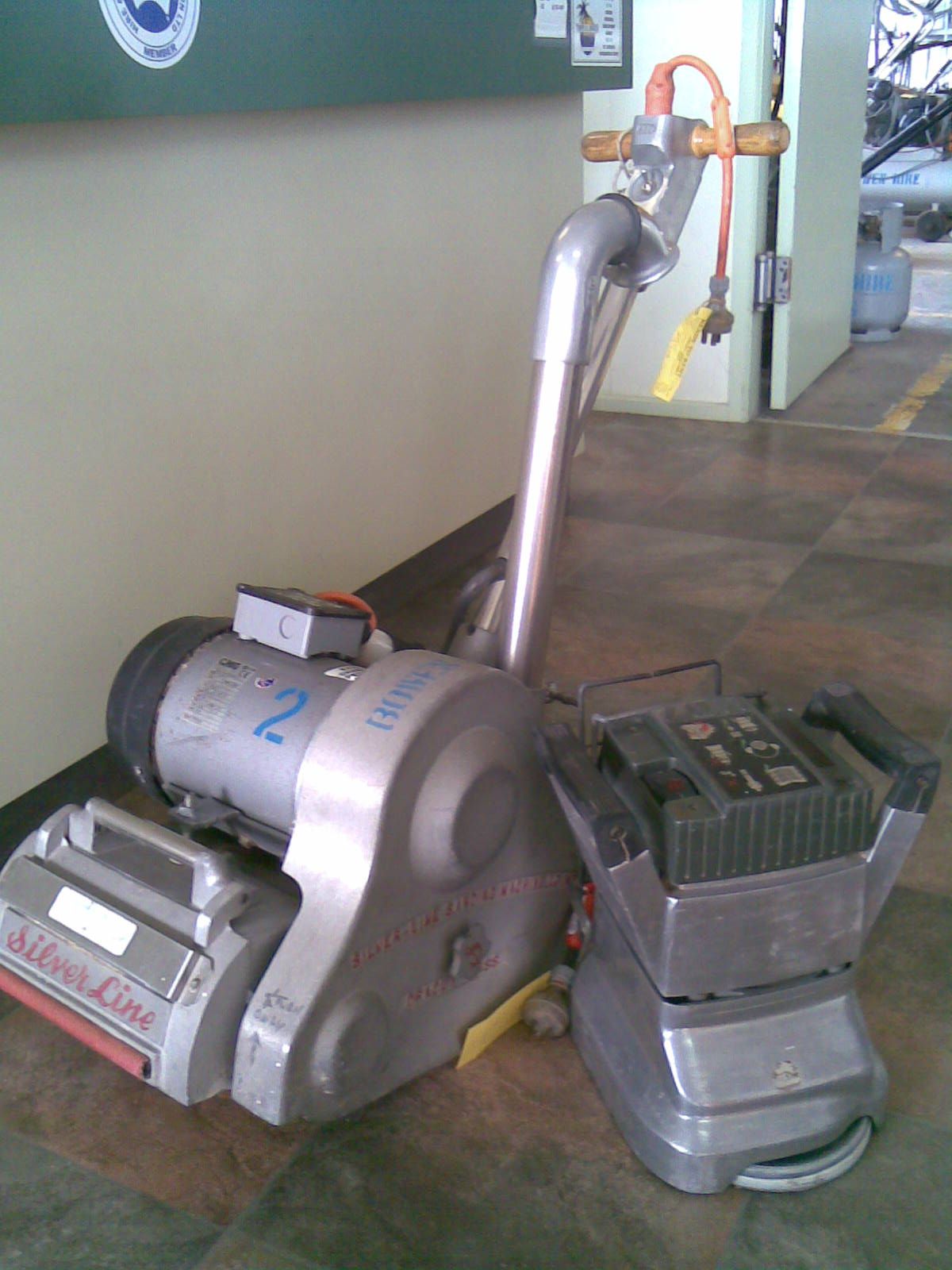 Floor buffer machine in a workshop, with a metal handle and attached dust collection unit.