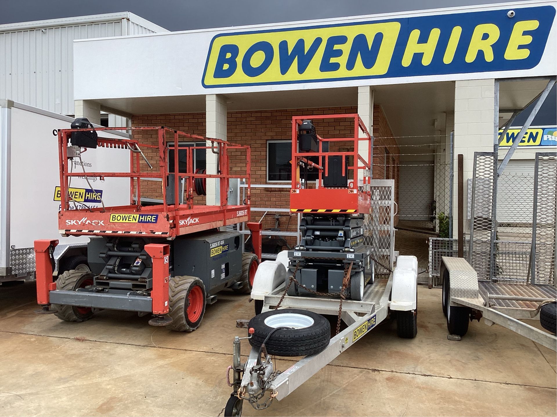 Two Red Boom Lifts and a Trailer Parked Outside Bowen Hire Storefront — Bowen Hire in Bowen, QLD