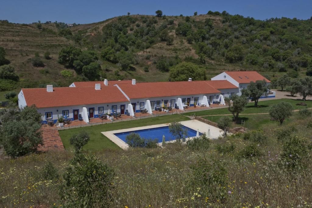 Um prédio comprido e branco com telhado de telhas vermelhas fica ao lado de uma piscina azul, cercado por colinas verdes.