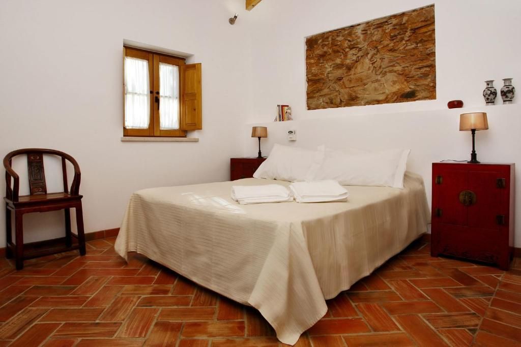 Quarto com cama de casal, duas mesas de cabeceira, uma cadeira e uma janela. Piso de azulejo marrom, paredes brancas.