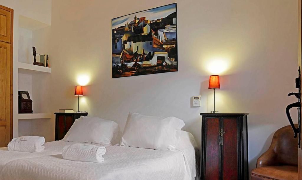 Quarto com roupa de cama branca, mesas de cabeceira, luminárias, obras de arte e uma poltrona de couro.