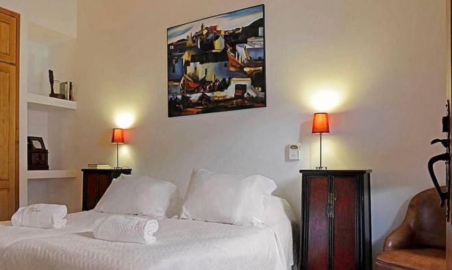 Quarto com roupa de cama branca, mesas de cabeceira, luminárias, obras de arte e uma poltrona de couro.