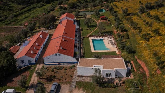 Vista aérea de edifícios brancos com telhados laranja, piscina, jardim verde e colinas circundantes.