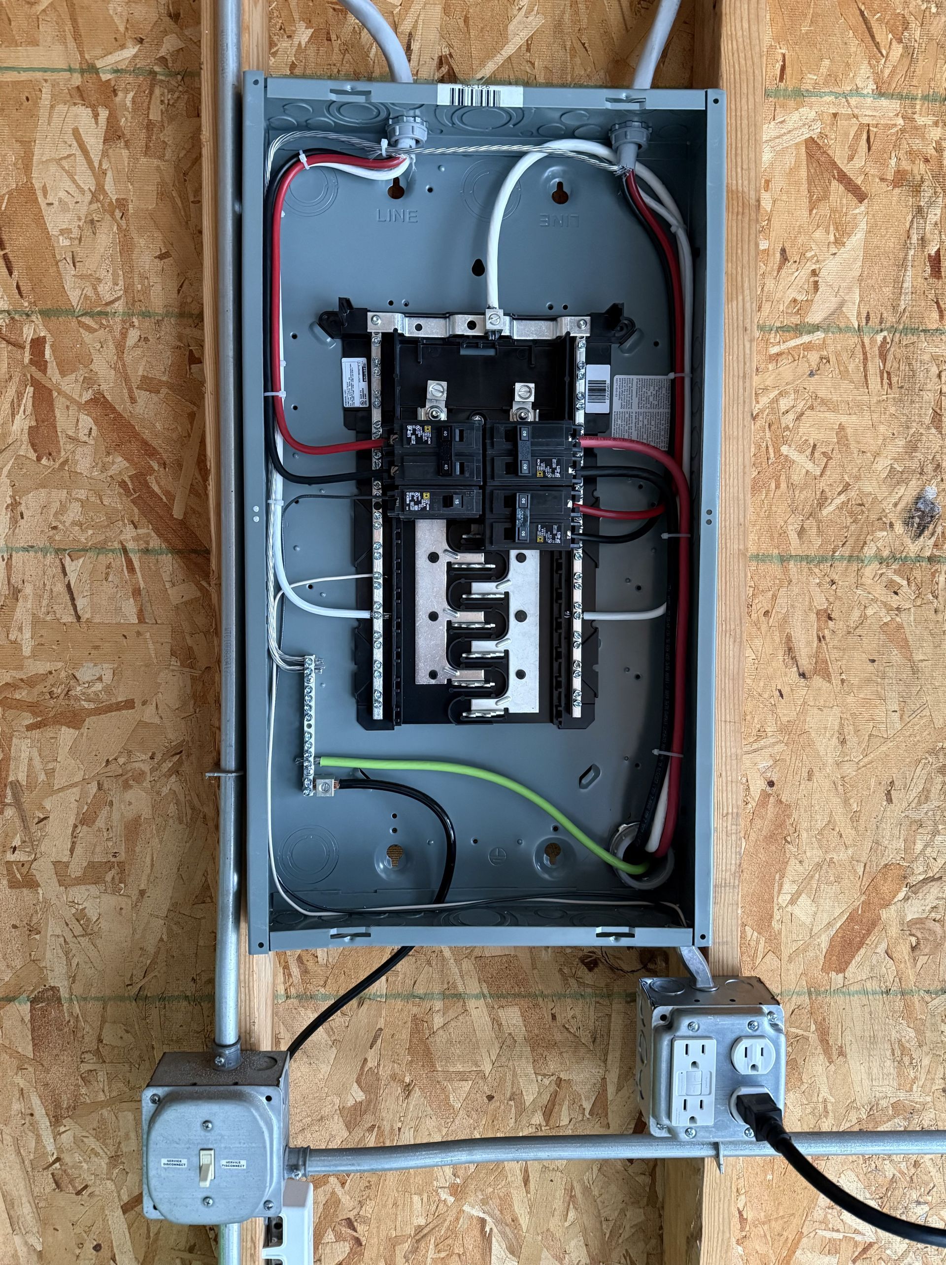 Electrical panel with wiring on a wooden wall. Gray box, black, red, white, and green wires.