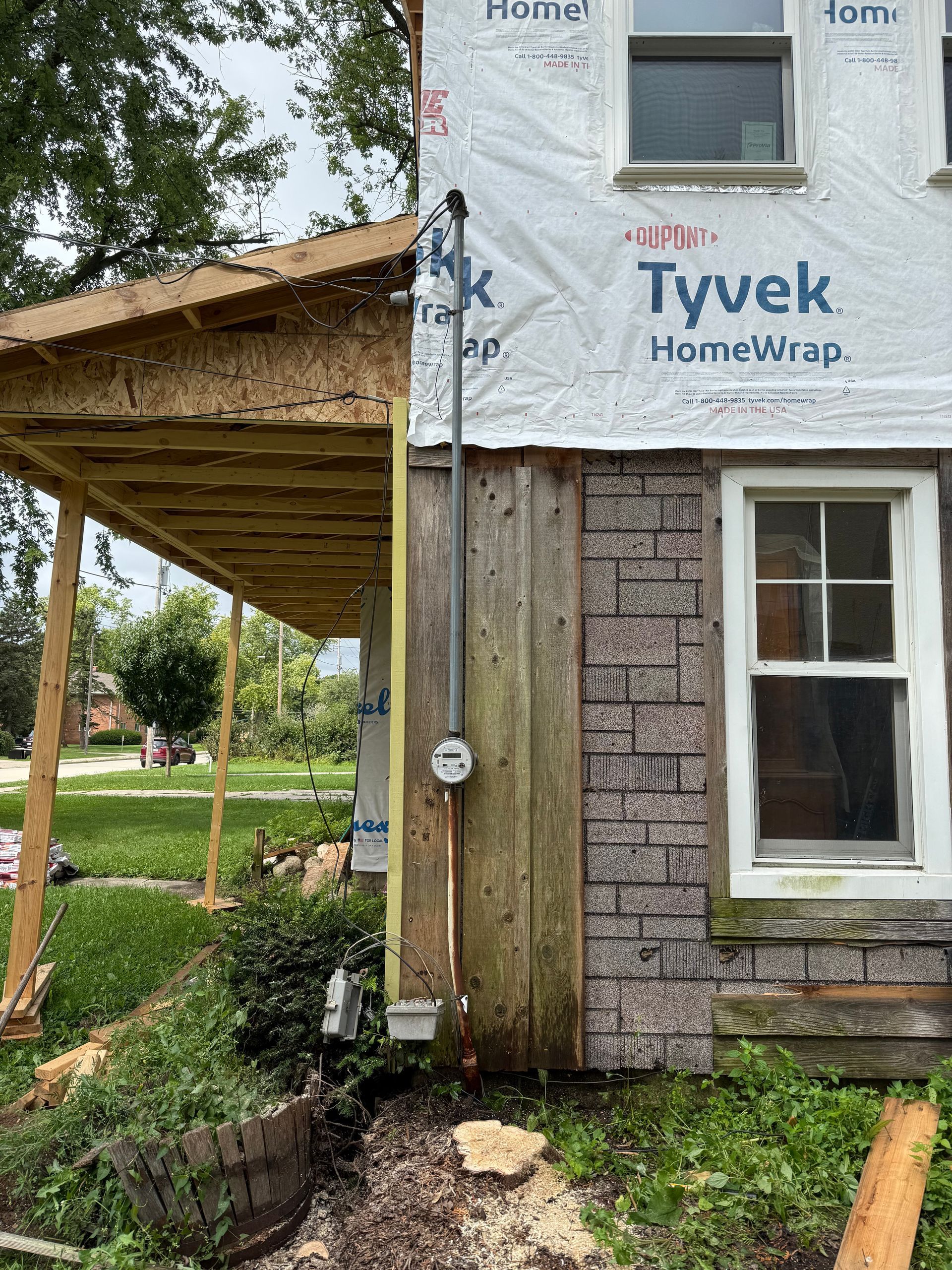 Electrical meter on a house exterior with a new covered porch under construction. Green grass, trees, and bushes in yard.