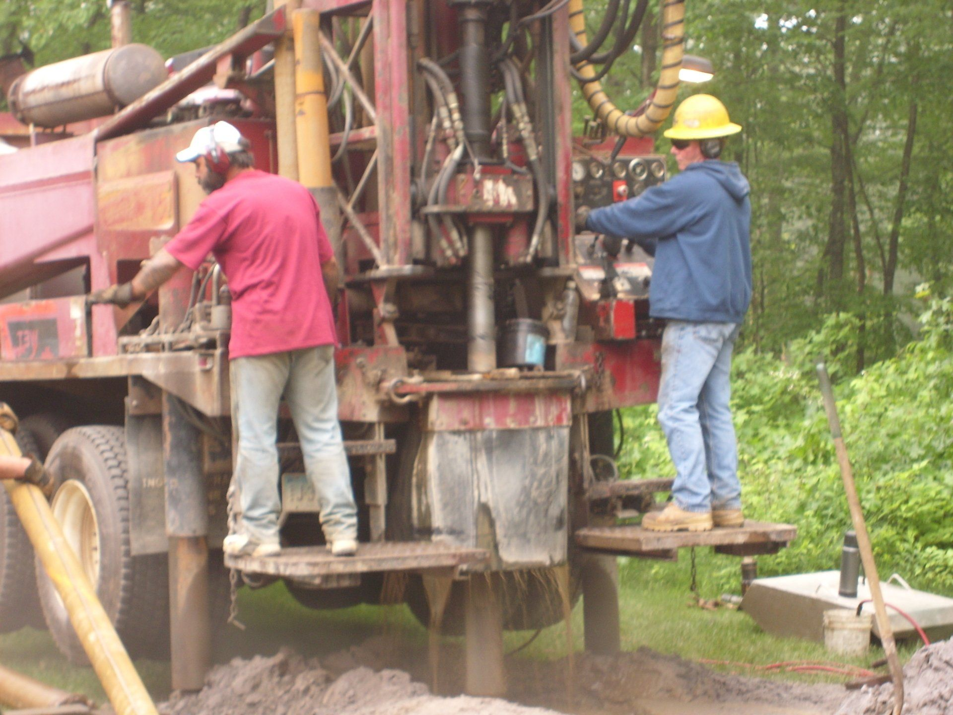 Two workers operating a drilling rig, outdoors. One wears a hard hat and blue jacket, the other a red shirt. Muddy ground.