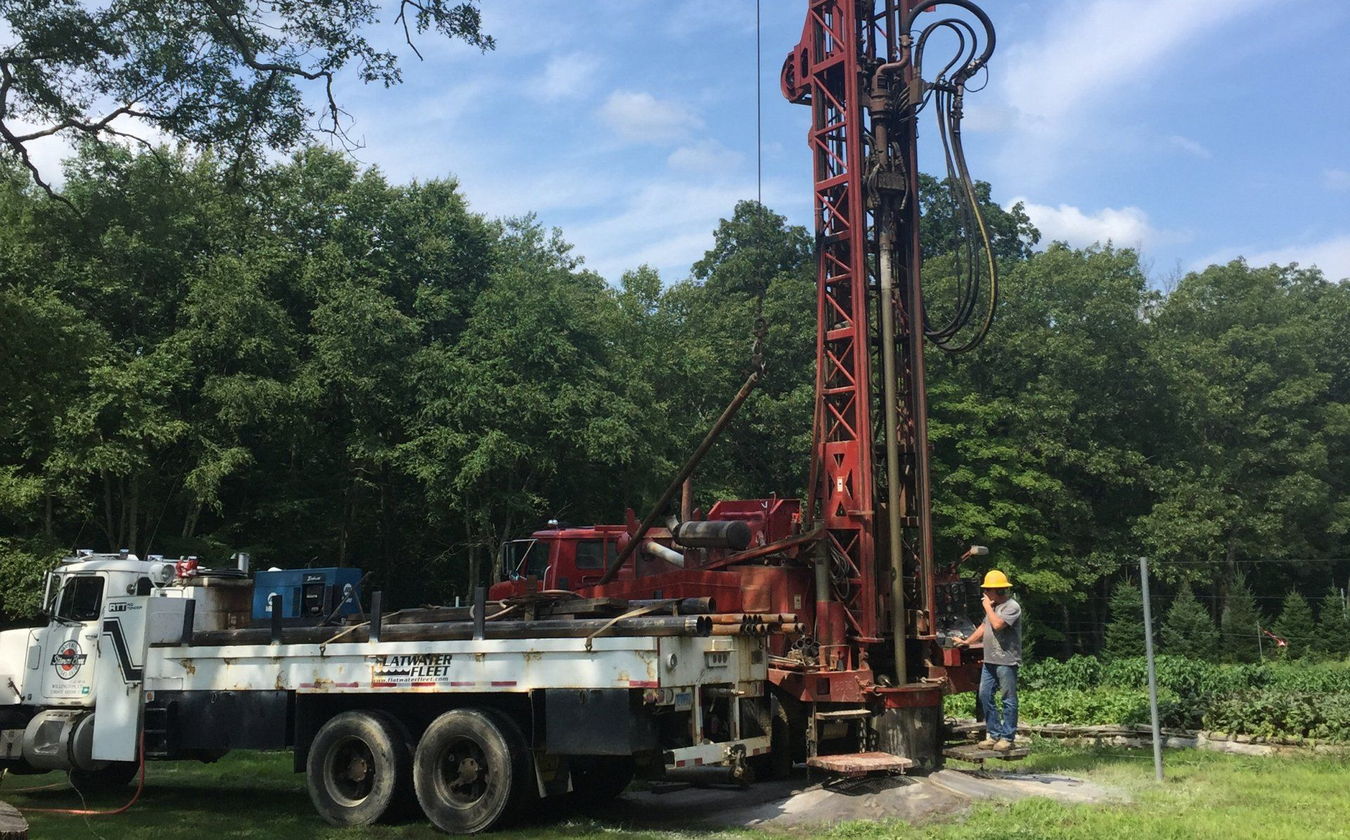 Well drilling rig on a truck, with a person operating it outdoors near trees.