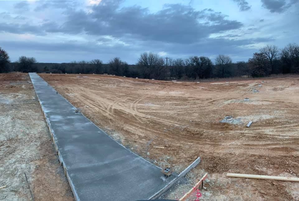 Concrete sidewalk under construction across a cleared dirt lot at dusk, with bare trees in the background