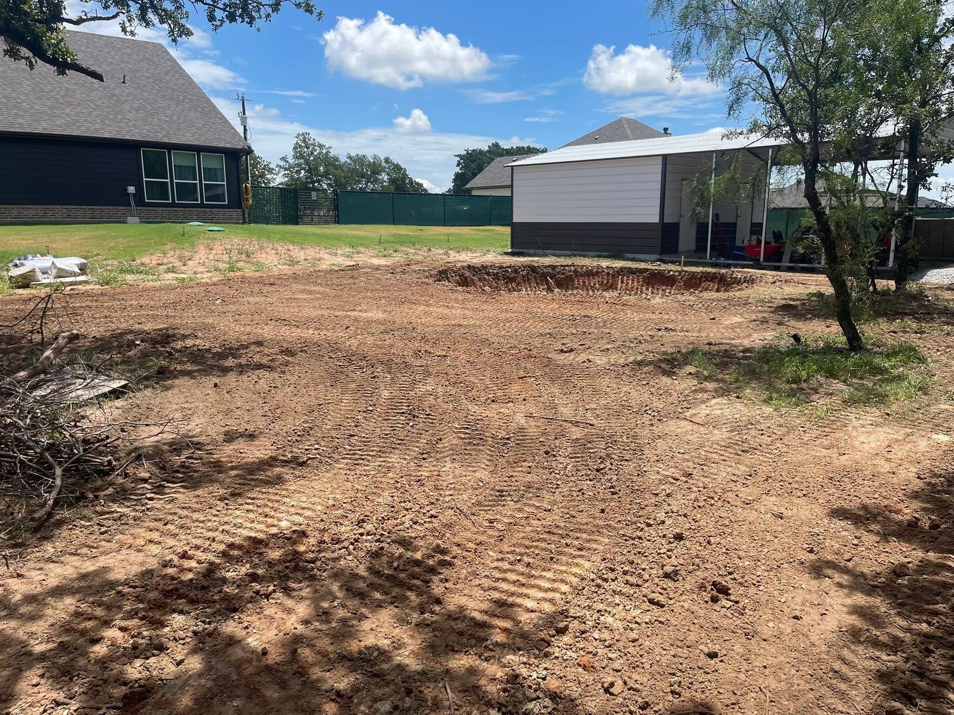Bare dirt lot beside homes and a detached garage under a sunny blue sky