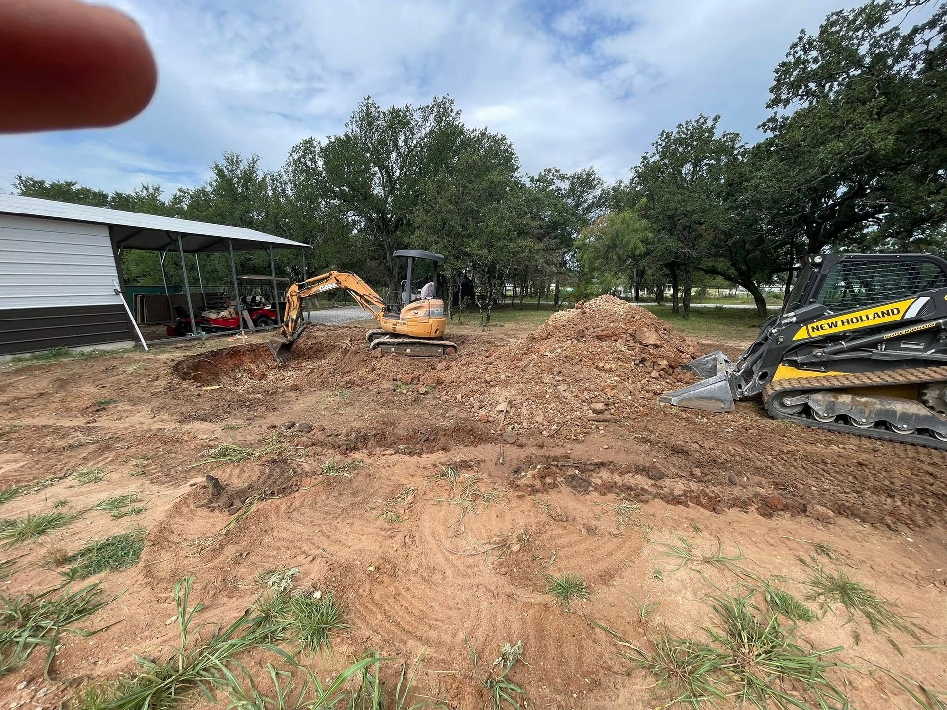 Excavators grading a dirt lot beside a shed and trees under a cloudy sky.