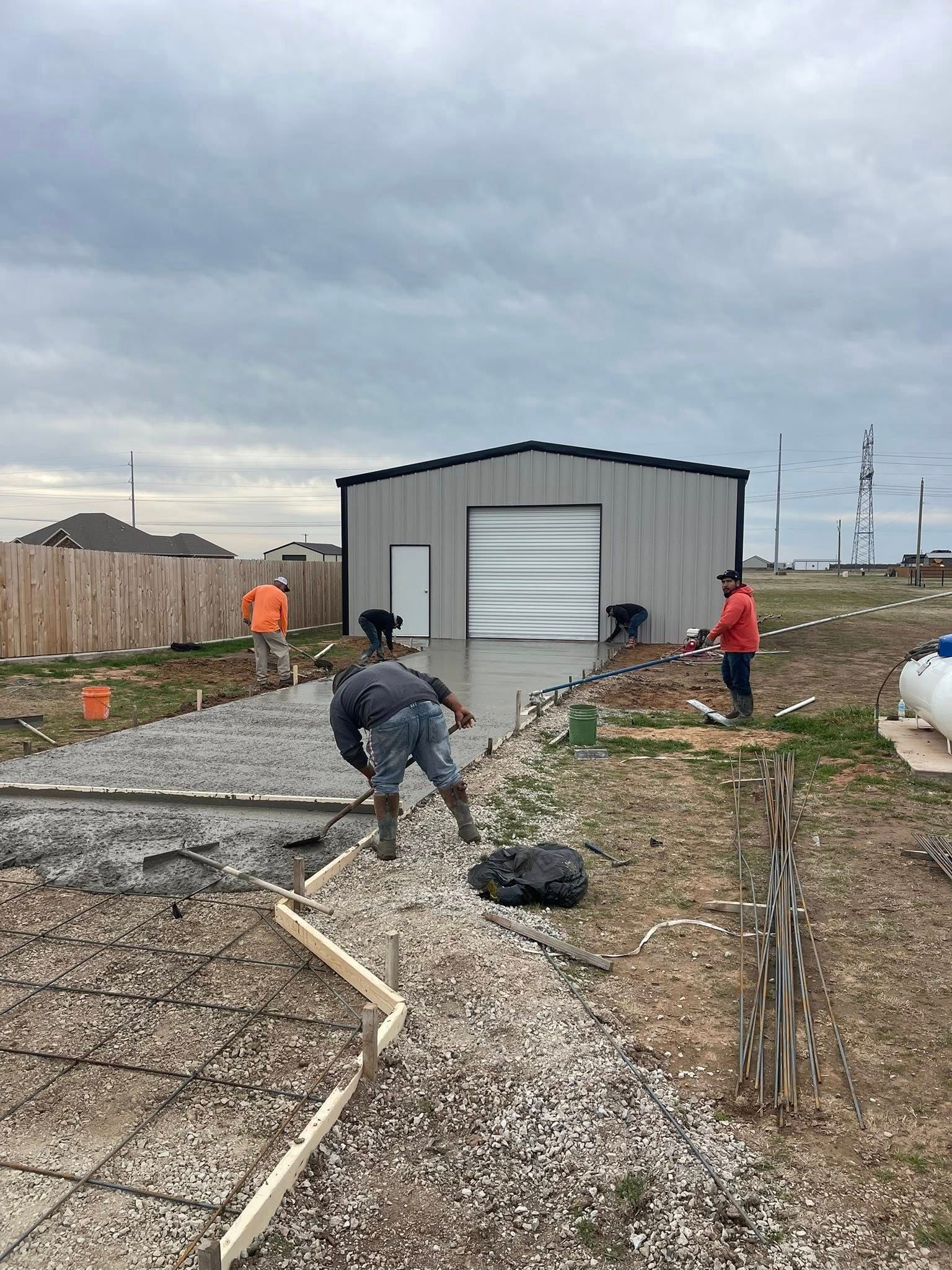 Two workers lay gravel by a small gray shed at a construction site under cloudy skies.
