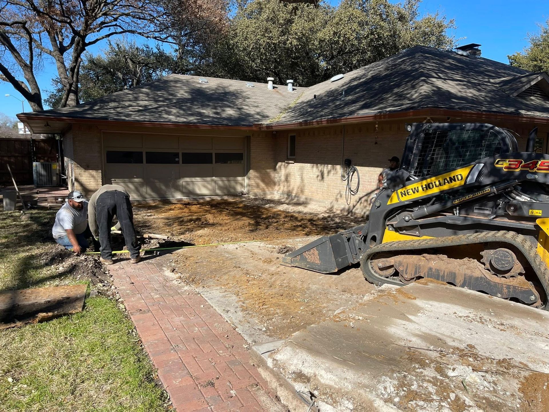 Worker using a jackhammer on a driveway beside a beige house with a yellow excavator nearby