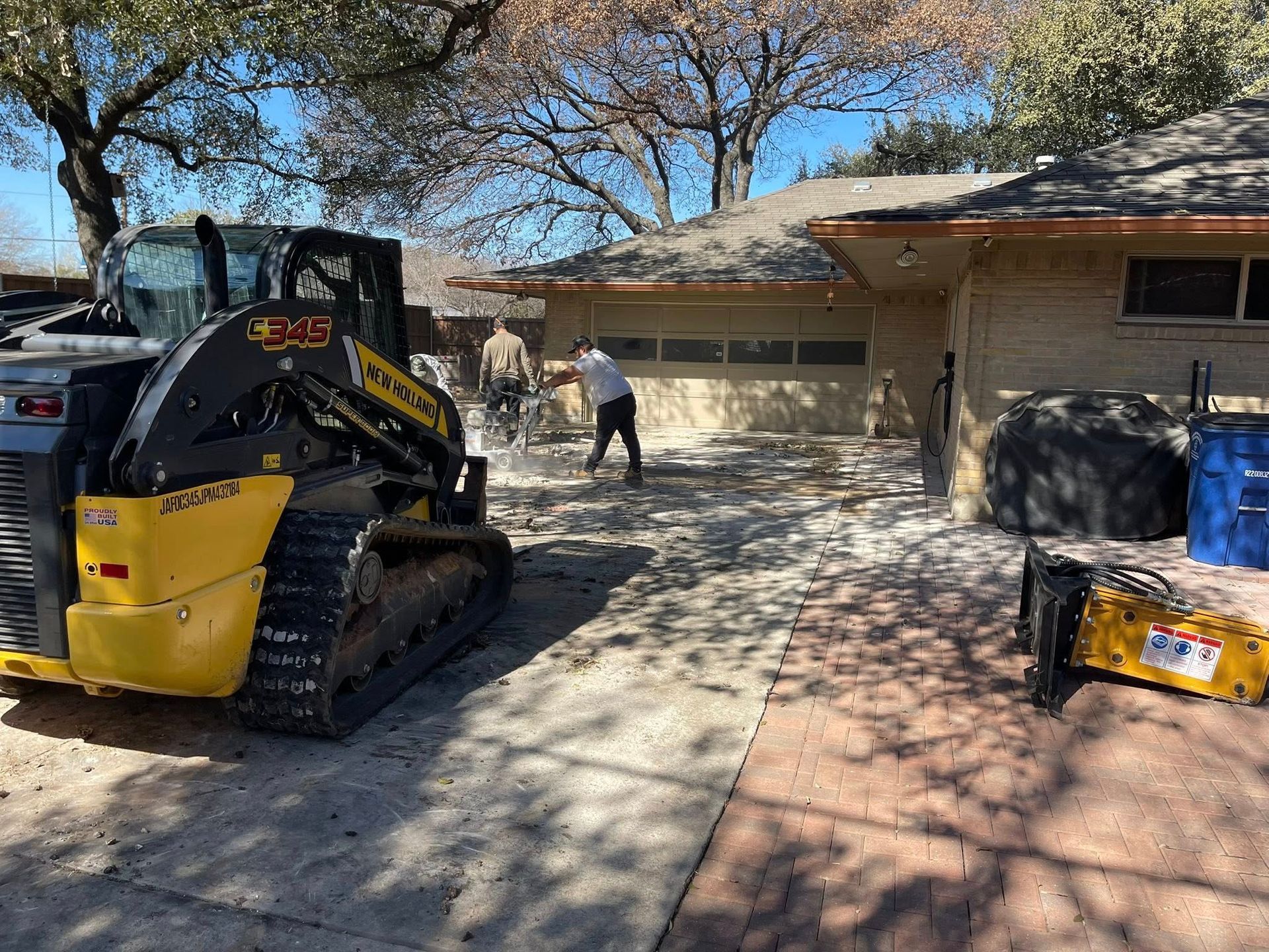 Yellow skid-steer in front of a house as workers clear debris from a driveway and yard