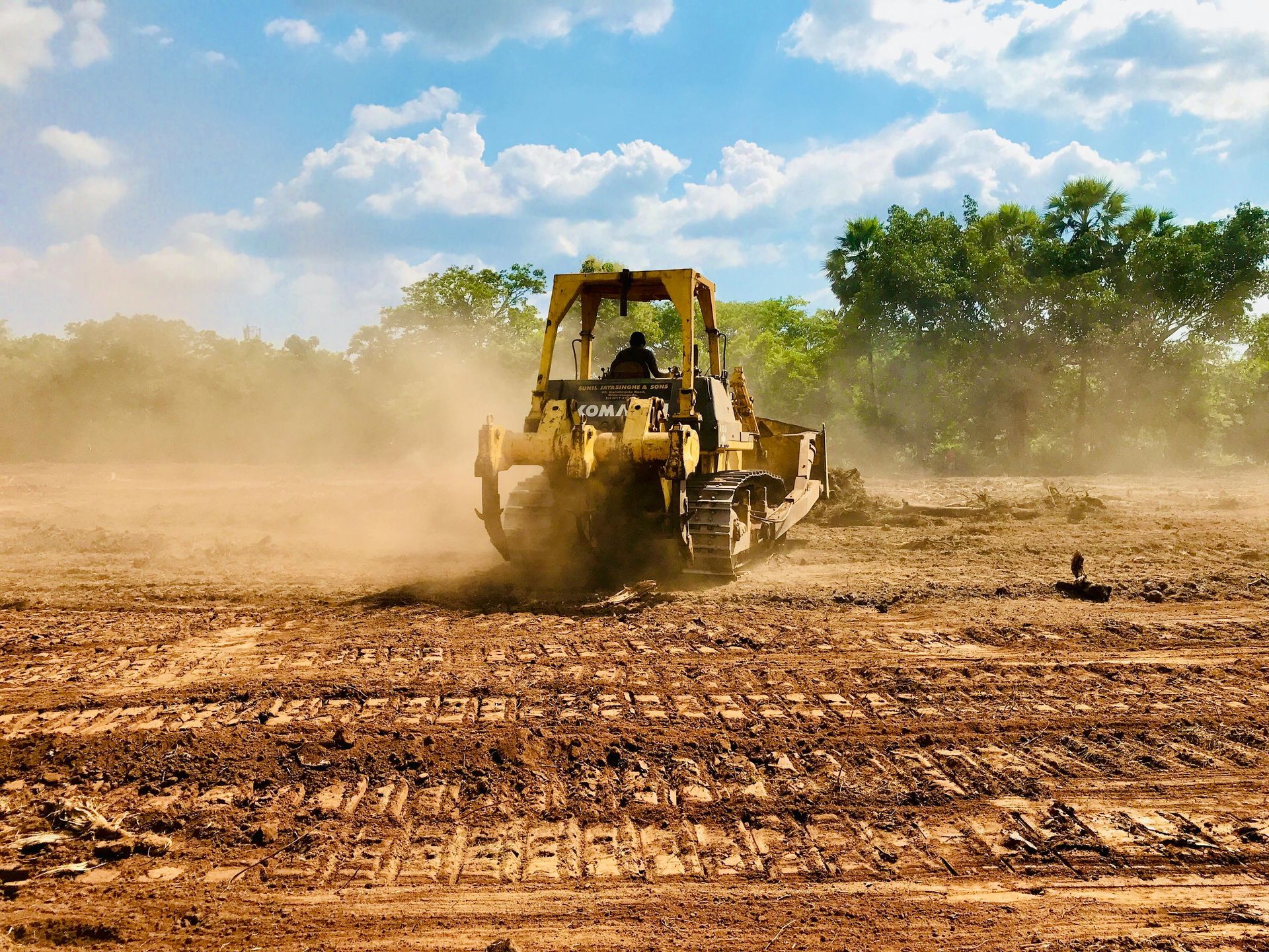 Yellow backhoe plowing a dusty field under a blue sky, with trees in the background