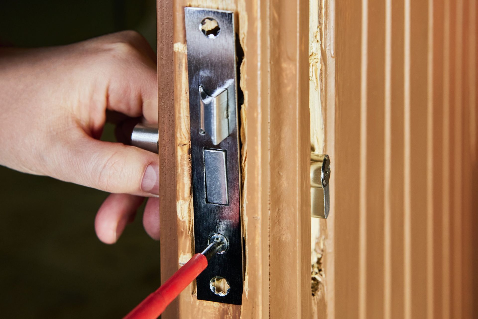 Hand opening a door latch with a red screwdriver on a wooden door frame